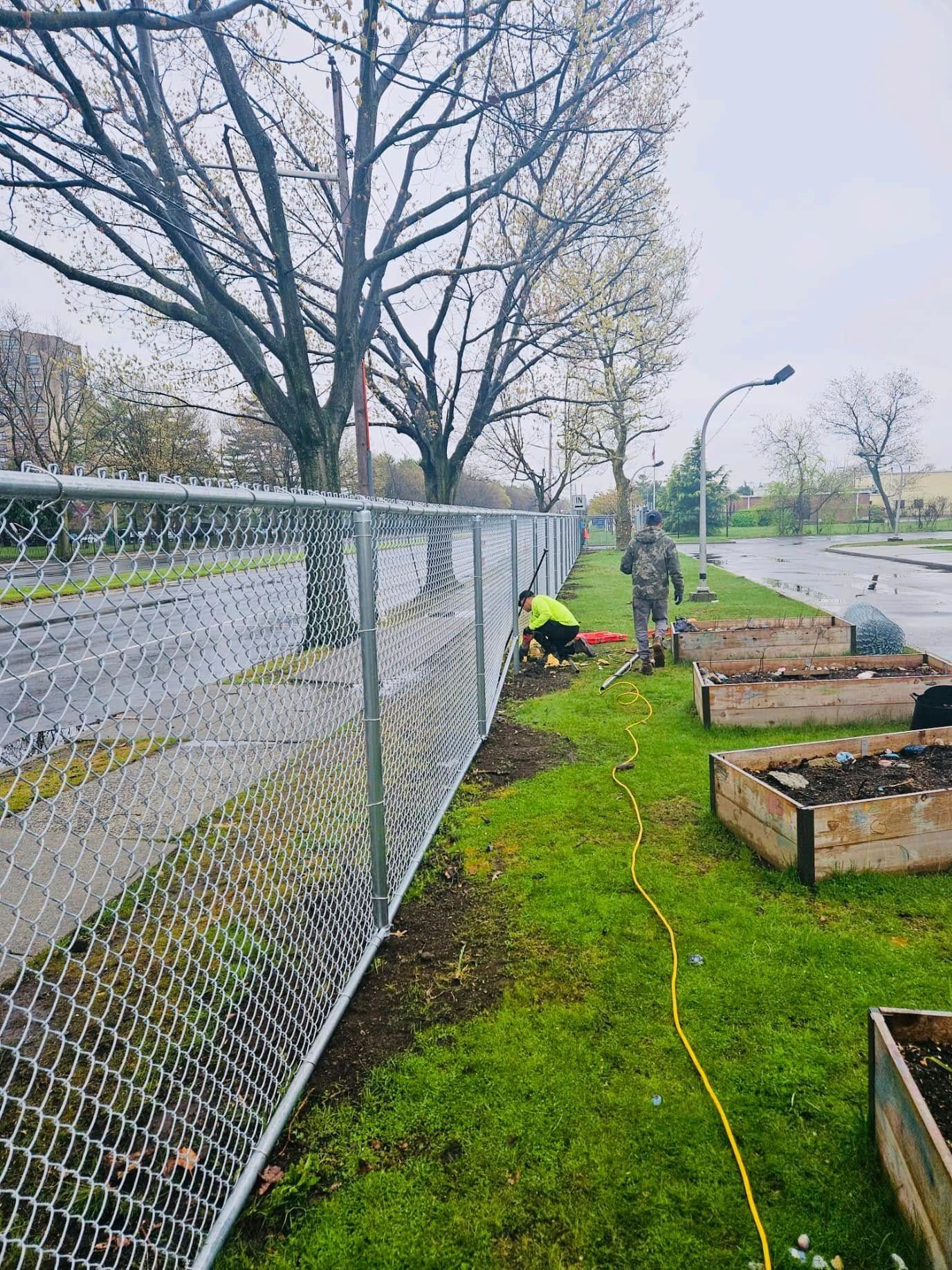 Workers tending raised garden beds next to a chain-link fence, under a cloudy sky.