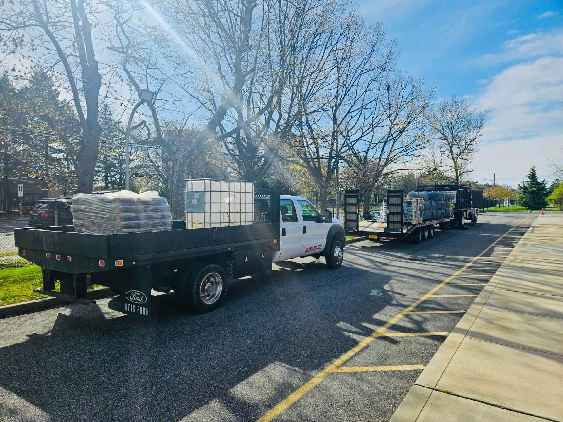 Two trucks hauling materials on a paved lot with trees in the background. Bright sunlight.