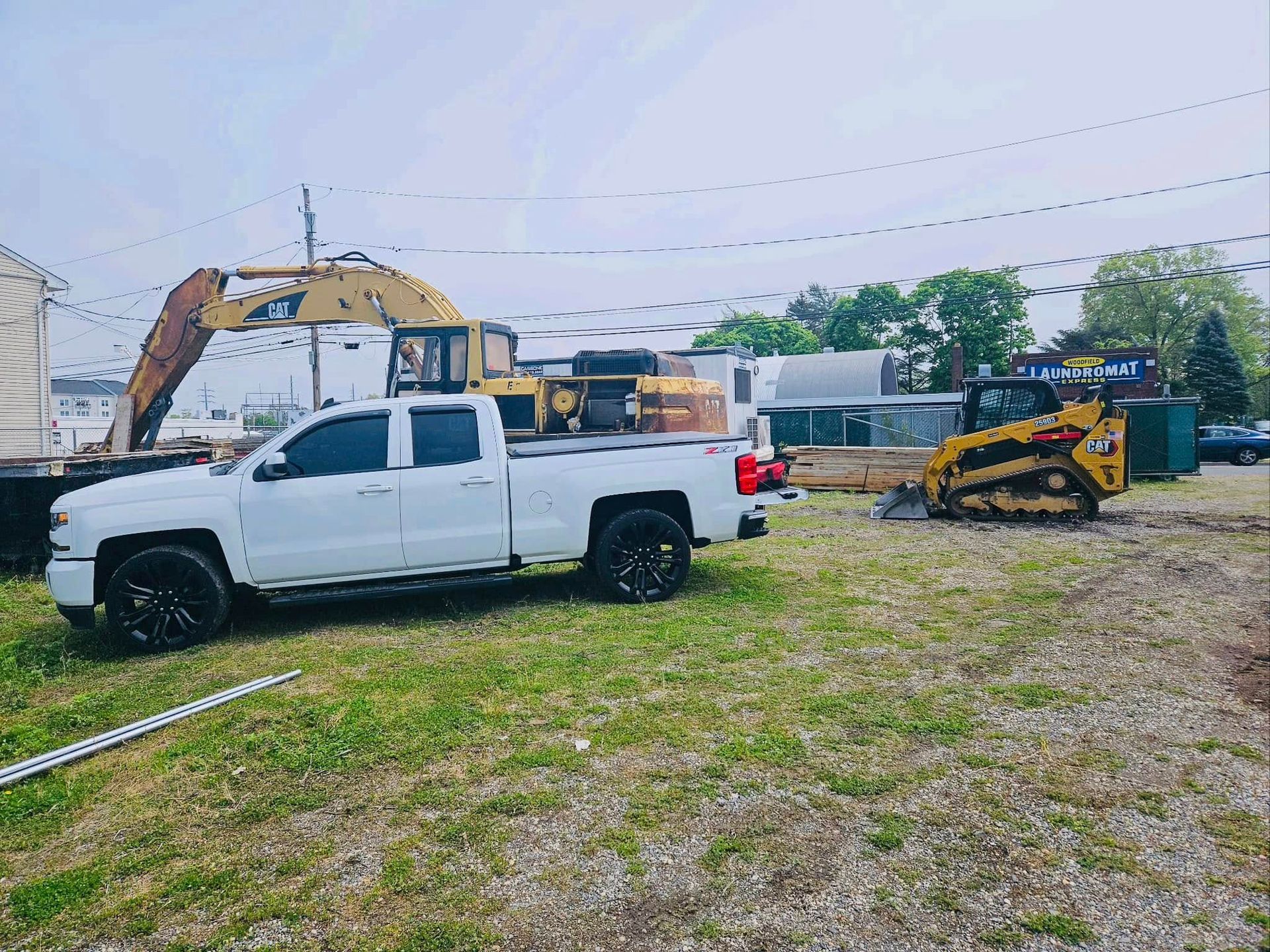 White pickup truck parked on a grassy lot with construction equipment in the background. Cloudy sky overhead.