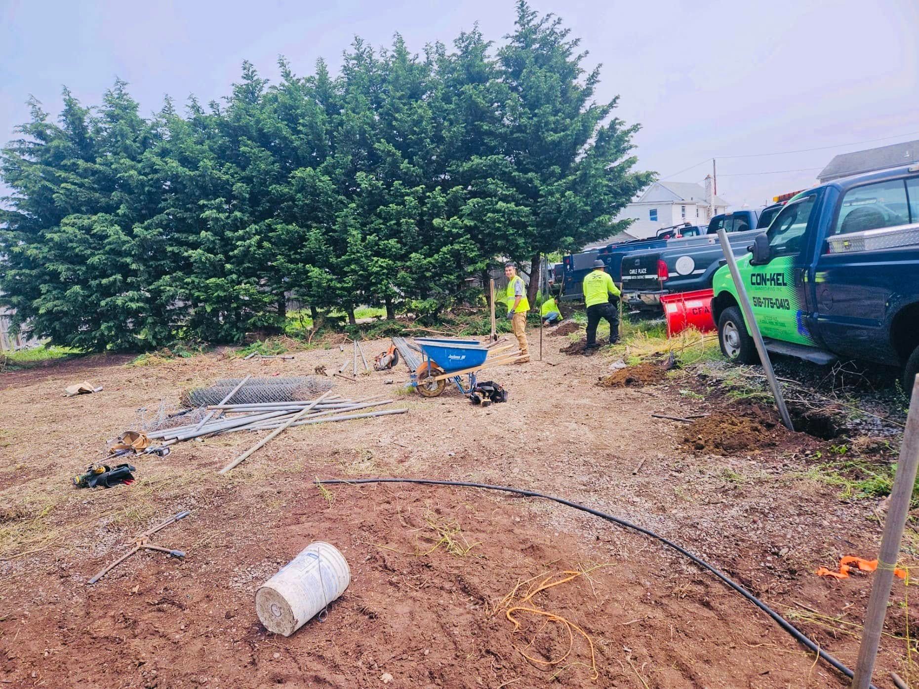 Construction workers near a row of trees with equipment, trucks, and materials on the ground.
