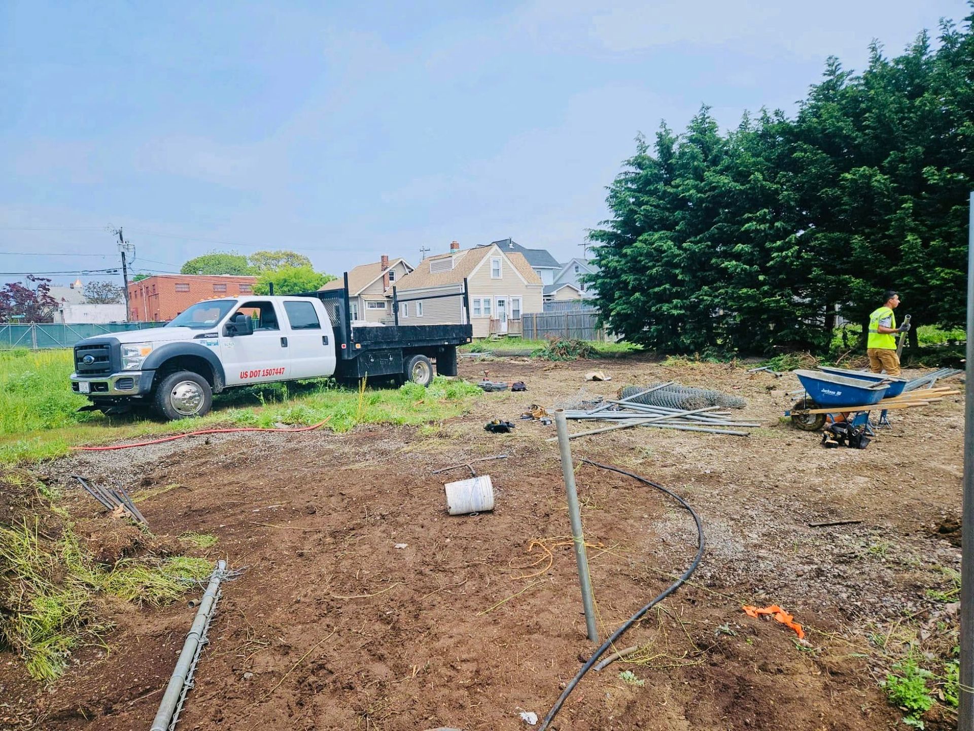 White truck parked on a dirt lot; construction site. A person in a high-vis vest stands near a wheelbarrow. Houses and trees in the background.