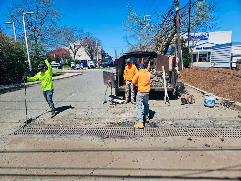 Road workers in orange and neon green clothing repair asphalt near a metal grate under a blue sky.