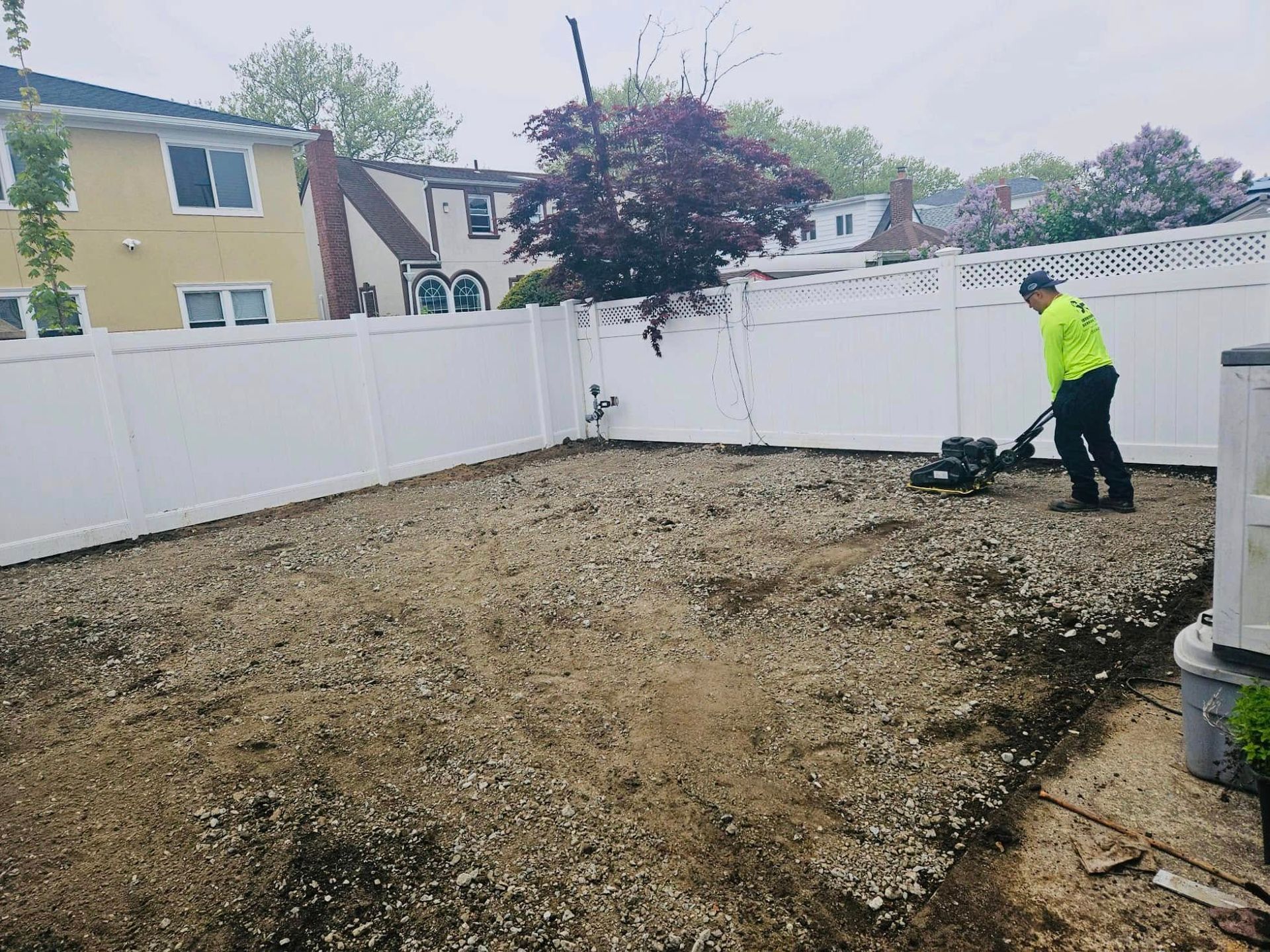 Person operating a machine in a yard with a white fence. The yard is covered in gravel and dirt.