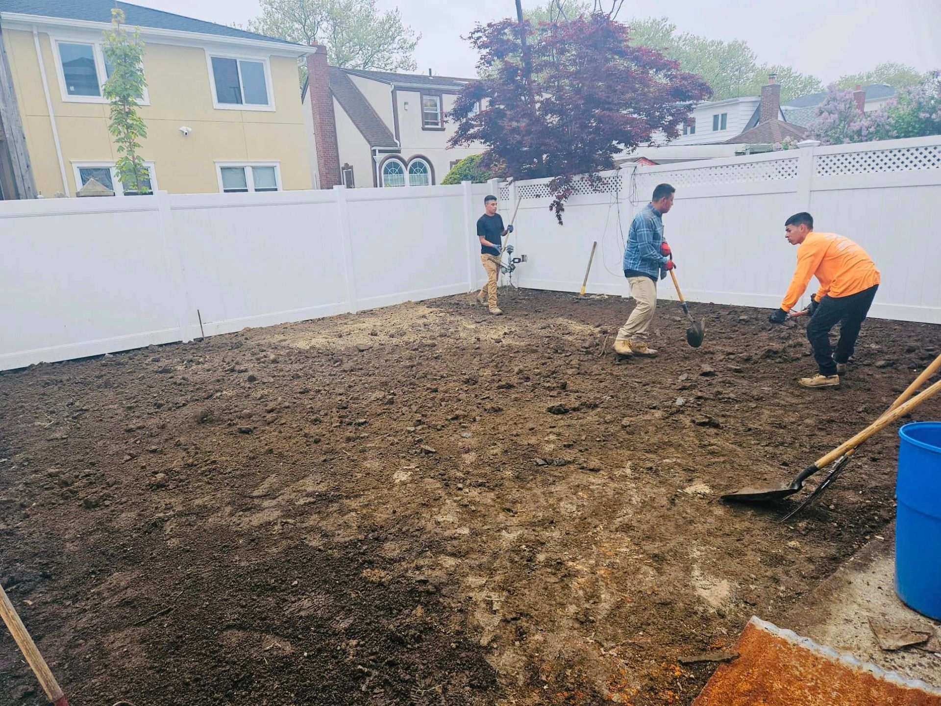 Three people digging in a backyard, preparing soil for planting, with a white fence in the background.