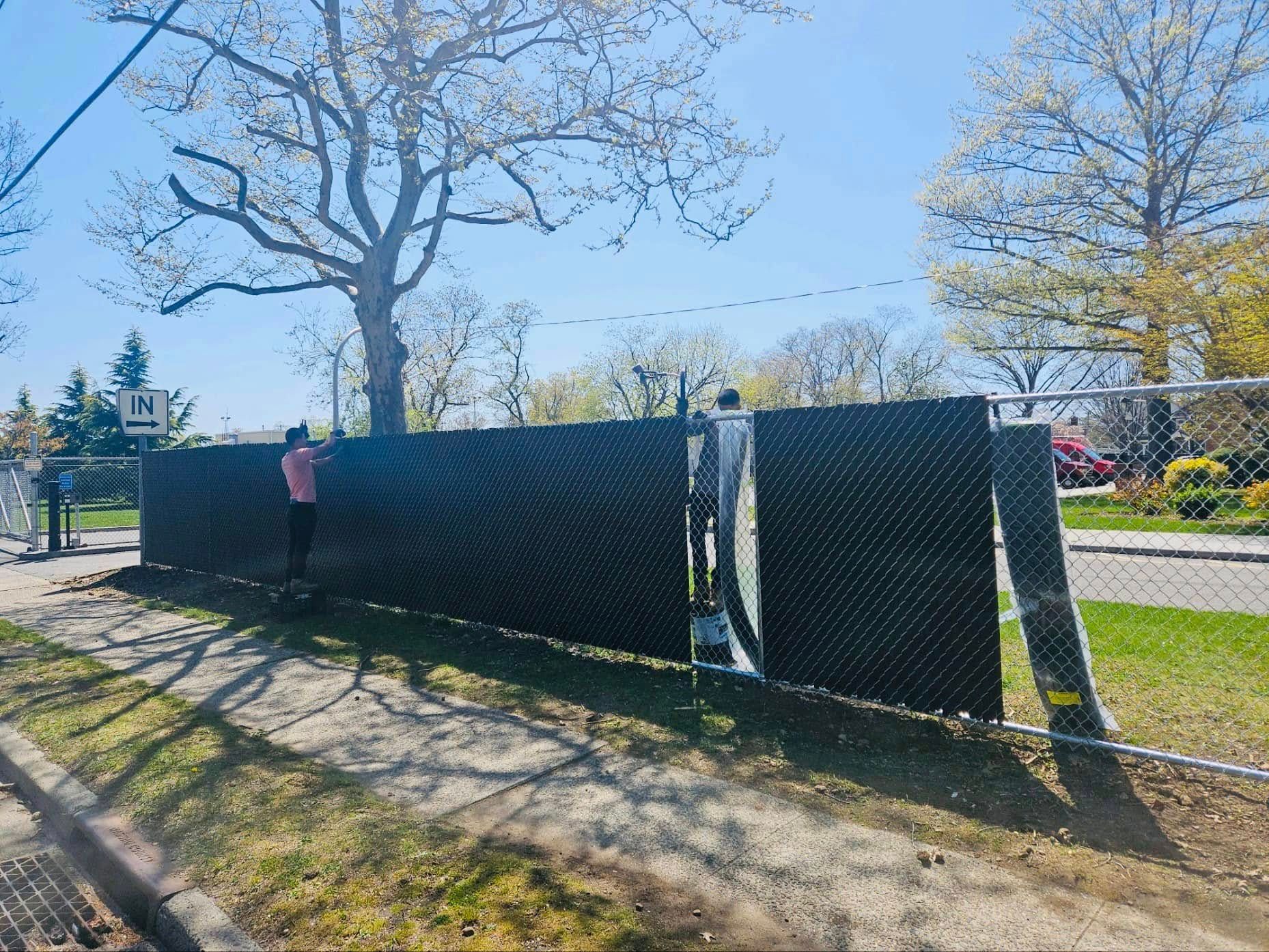 Person attaching a black privacy screen to a chain-link fence on a sunny day.