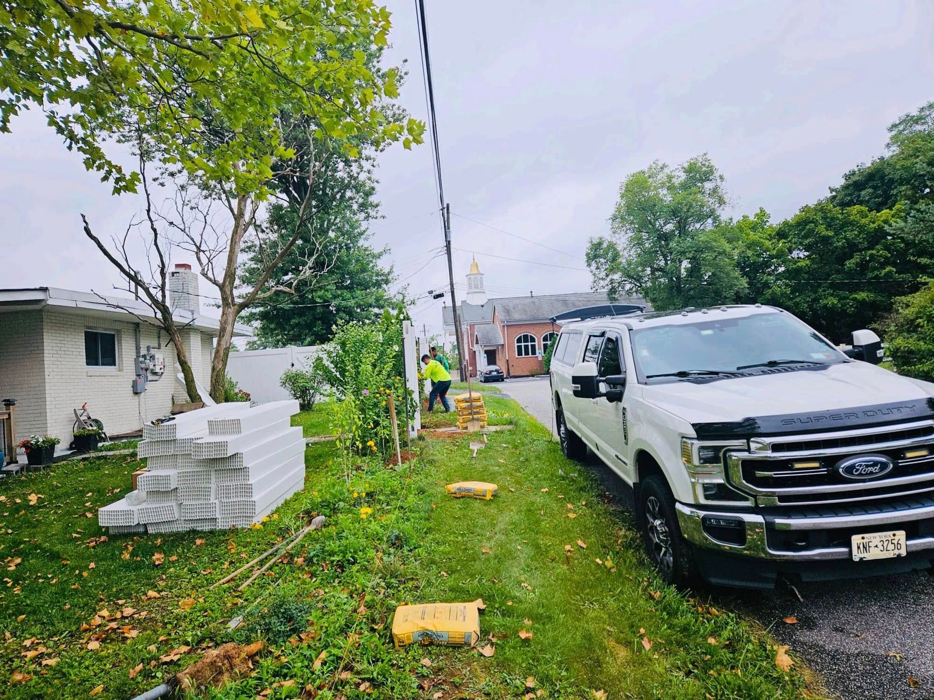 A worker near a white truck, green grass, and stacked blocks next to a house with power lines overhead.
