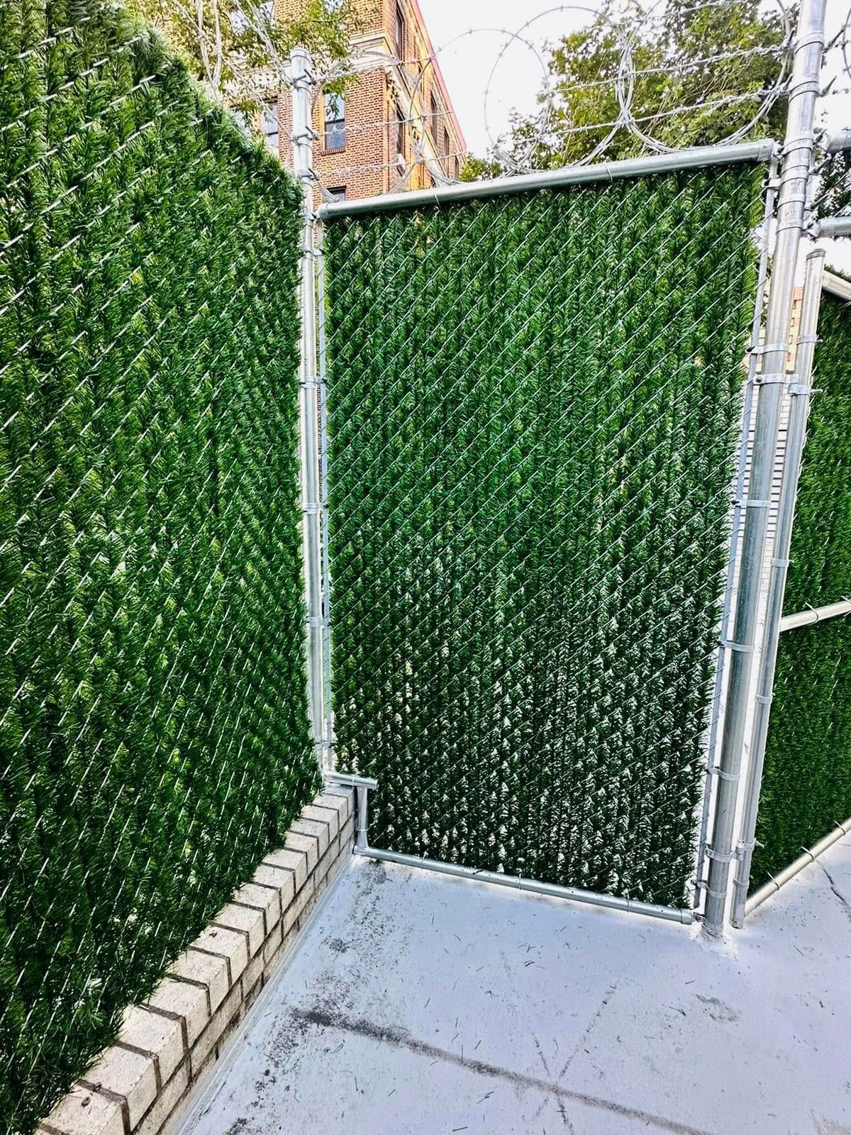 Green privacy fence made of chain link with faux foliage, beside a brick wall.