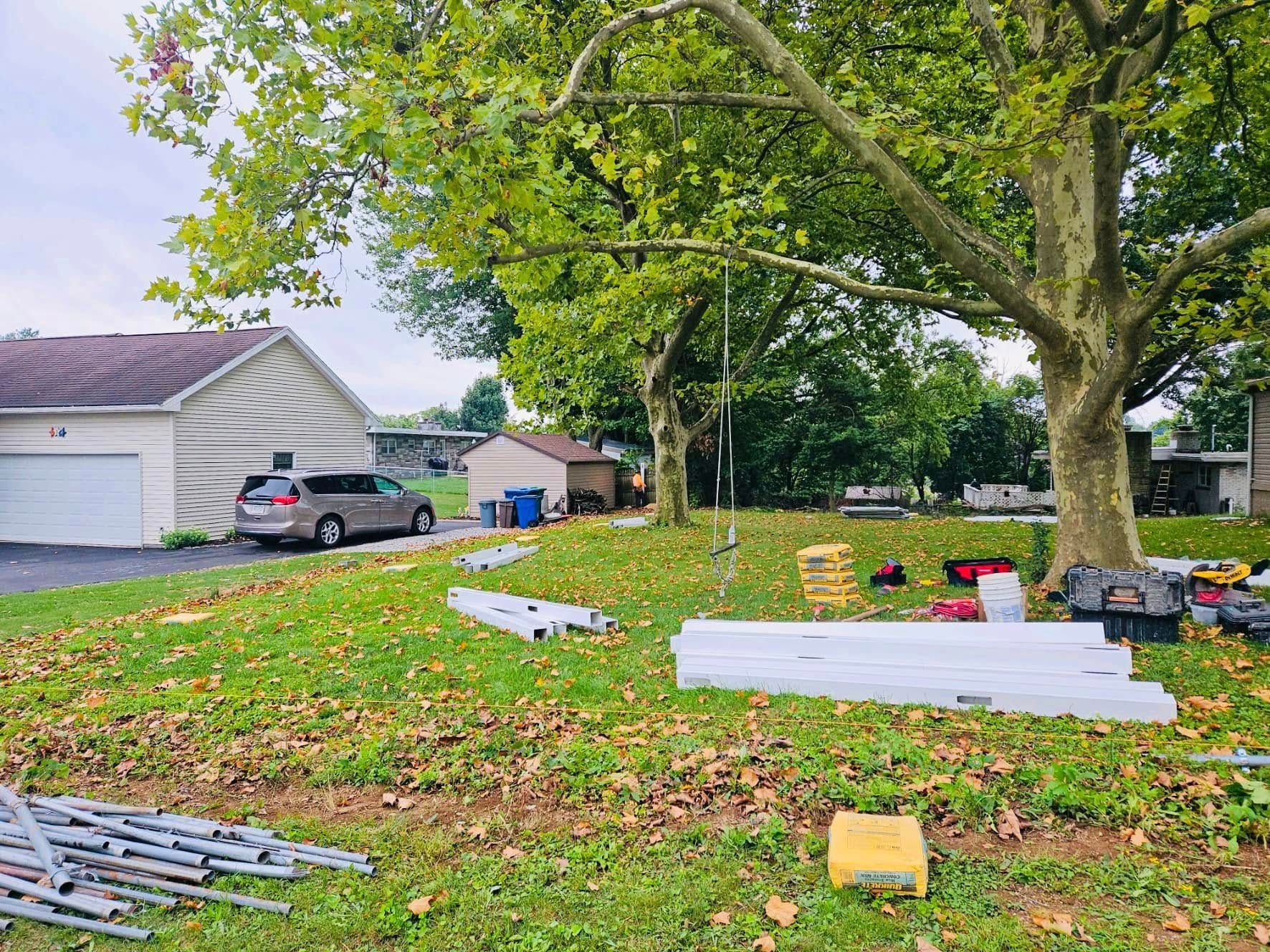 Backyard with a car parked near a garage. Construction materials scattered on the lawn under a large tree.