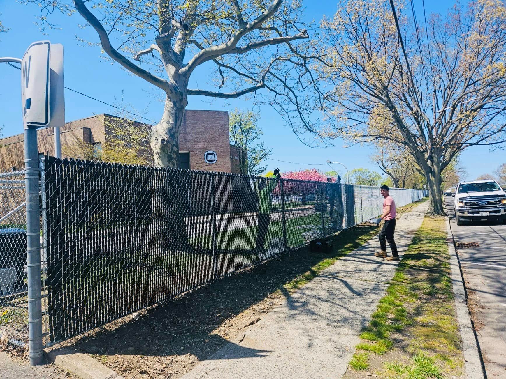 Black fence along a sidewalk, a person walking, car parked. Sunny day.