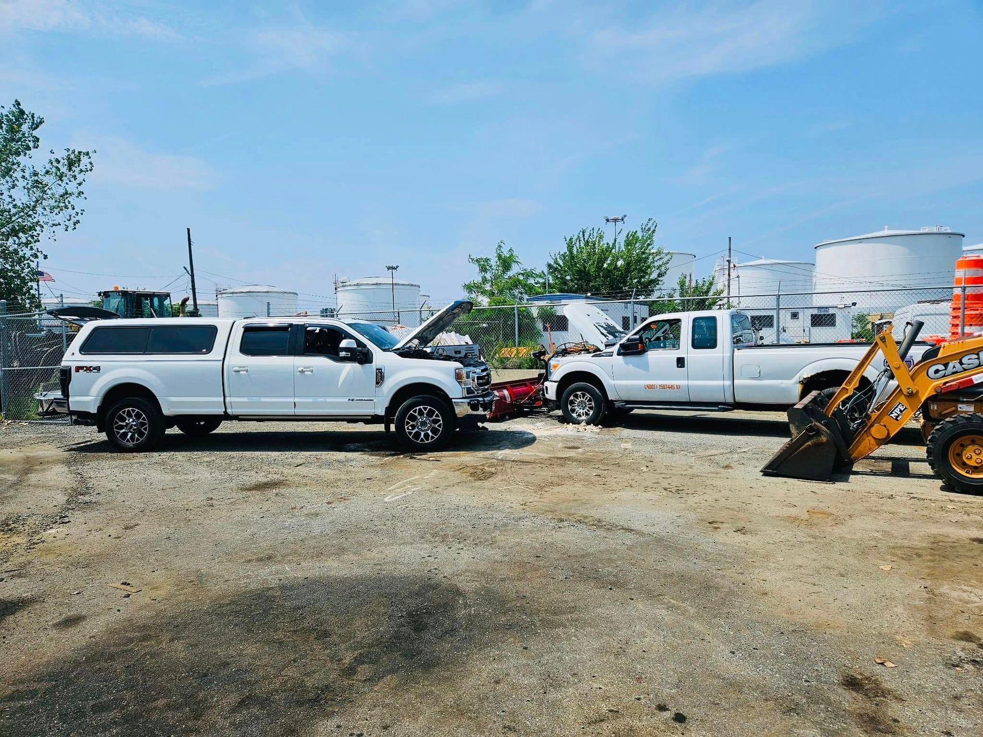 Two white pickup trucks with open hoods, connected with jumper cables, in a gravel lot with tanks and a backhoe.