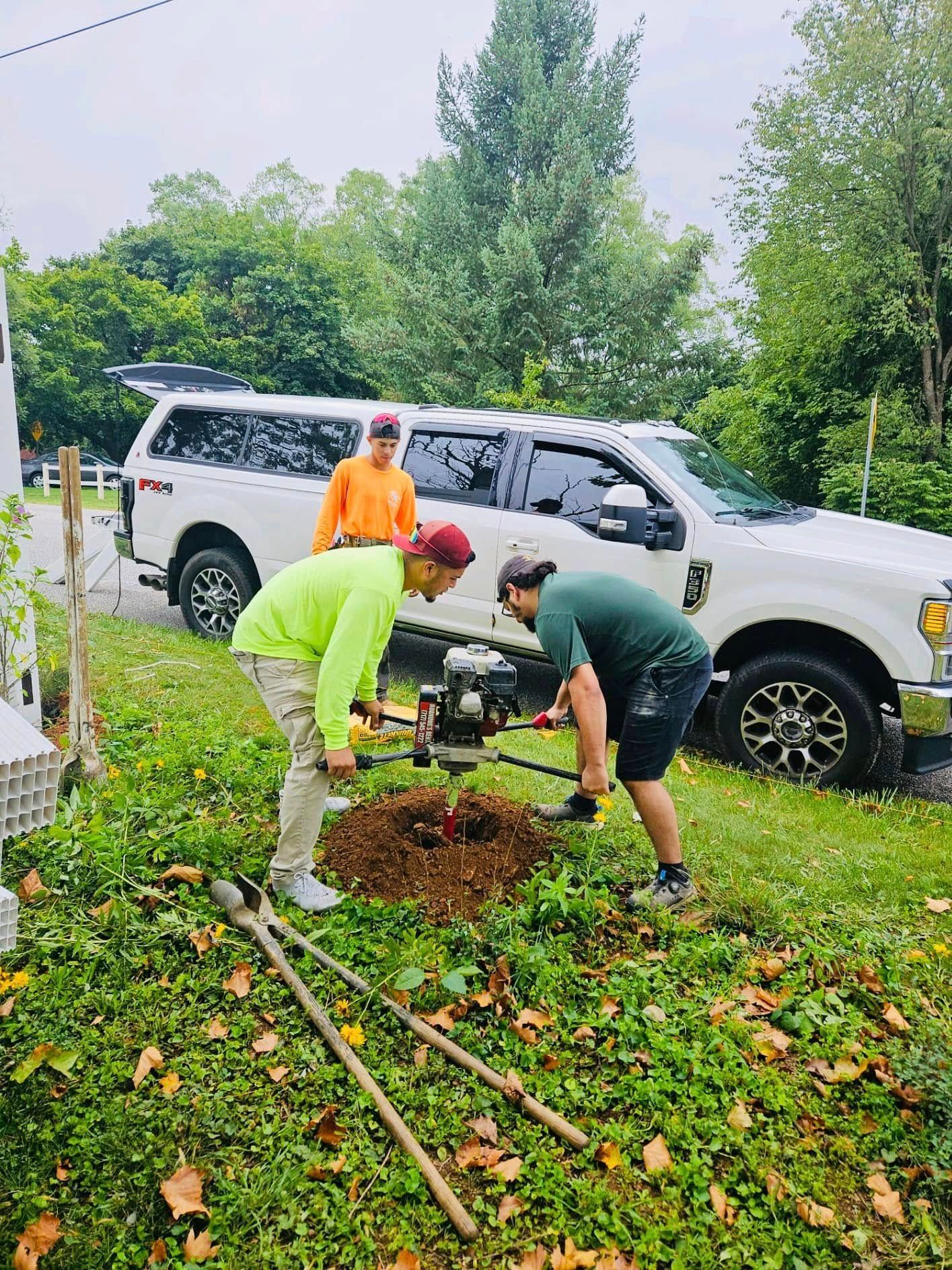 Three people planting a tree near a white pickup truck. One uses a power auger, two others watch.