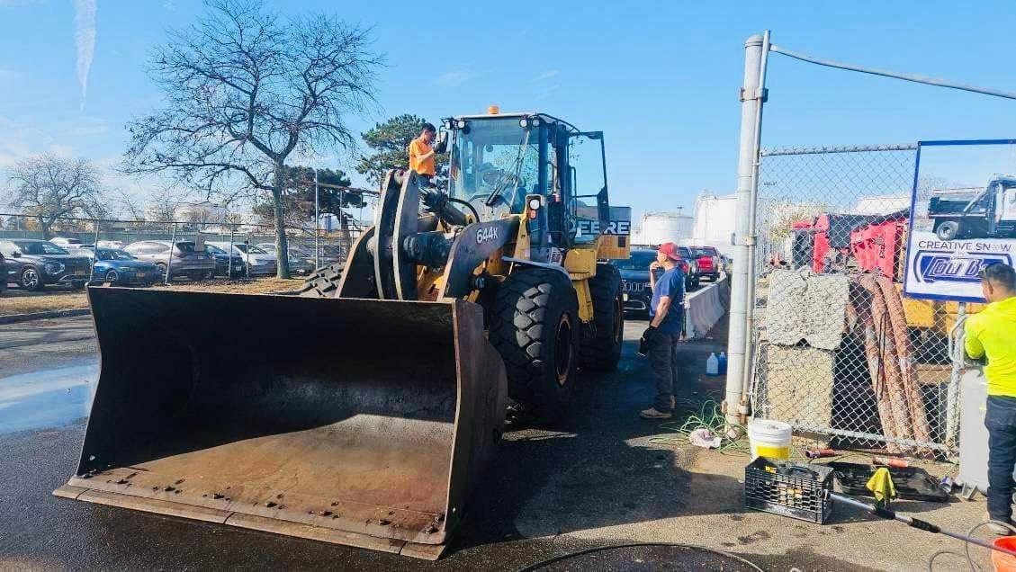 Large front-end loader with a blade parked near a fence, two people stand beside it on a sunny day.