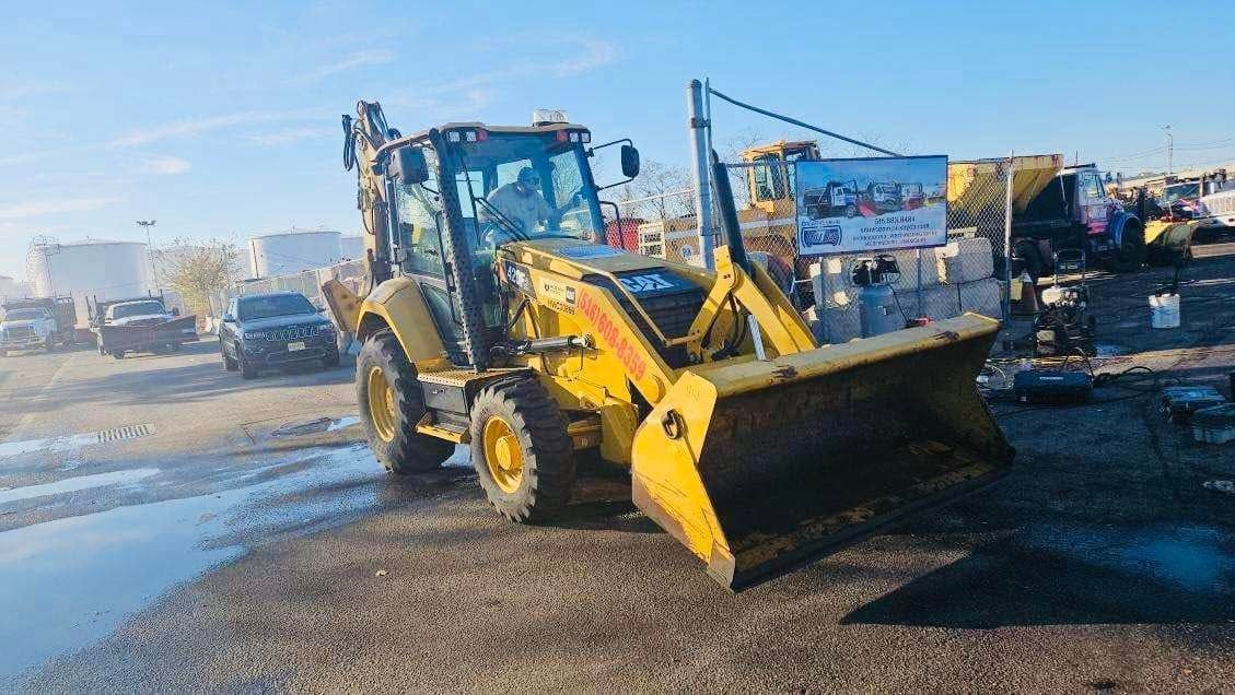 Yellow backhoe on a wet surface with buildings in the background. A sign is visible.
