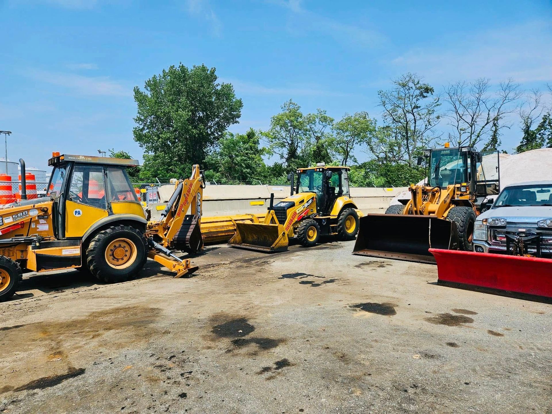 Yellow construction vehicles parked on pavement under a blue sky, near trees.