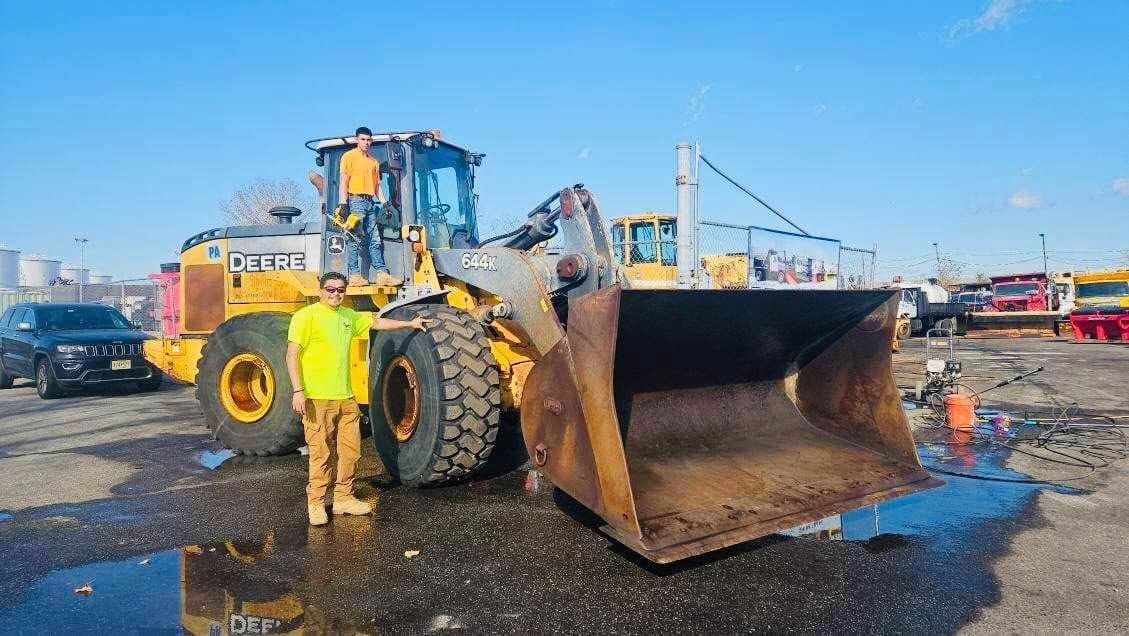 A man in a yellow vest stands beside a large front-end loader on a paved lot. Another person stands on the loader.