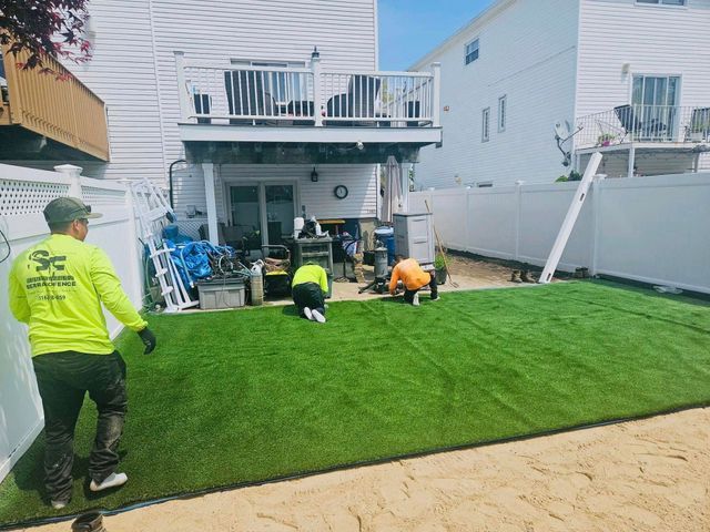 Workers installing artificial turf in a backyard beside a white house with a deck and patio.