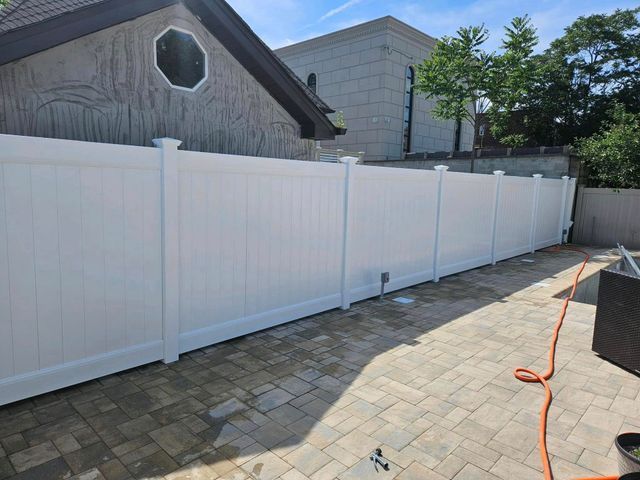 White vinyl fence along a paved patio beside a house, with an orange hose on the ground.