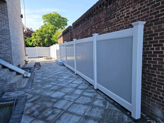 White vinyl privacy fence along a paved side yard beside a brick building on a sunny day