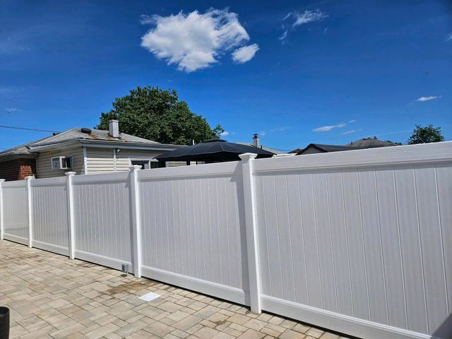 White vinyl fence along a paved driveway beside a house under a bright blue sky