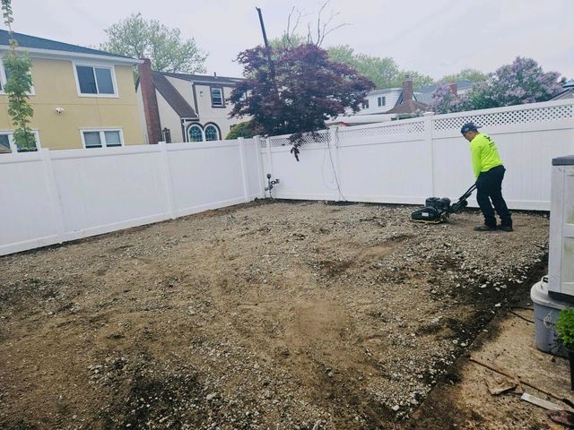 Worker leveling a dirt backyard inside a white-fenced yard, wearing a neon jacket.