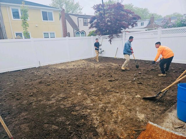 Workers spreading soil in a backyard beside a white fence and blue barrel