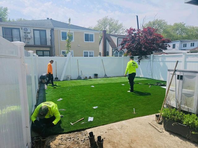 Workers installing turf in a backyard with white fencing and a two-story house in the background