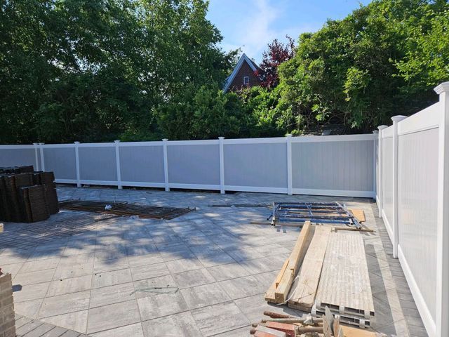 Backyard patio with gray pavers, white fence, stacked lumber, and trees beyond the yard