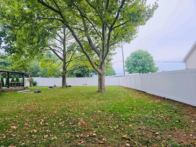 Large grassy backyard with a tree, white fence, and covered patio under a cloudy sky