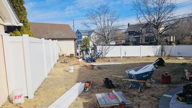 Backyard under construction with a white fence, dirt ground, tools, and a wheelbarrow.