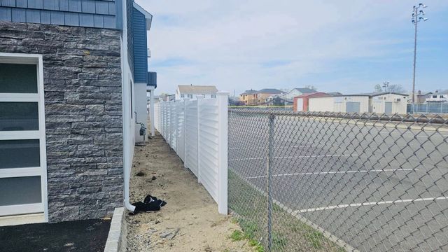 Residential side yard beside gray stone house with white fence and chain-link fence along a gravel path