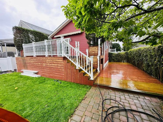 Red house with white porch railings and wooden deck, beside a green lawn and wet brick patio.