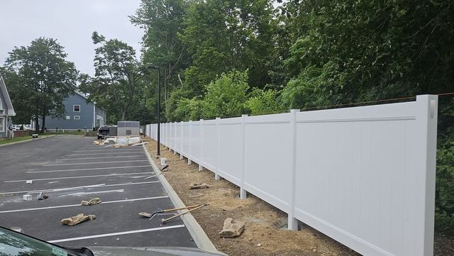 White vinyl fence along an empty parking lot beside trees and a house on a cloudy day