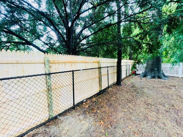Chain-link fence beside a beige wall under large trees, with a dirt path and a white gate in the distance