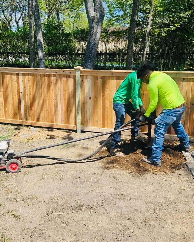 Two workers in green shirts clearing debris beside a wooden fence with a pressure washer hose.
