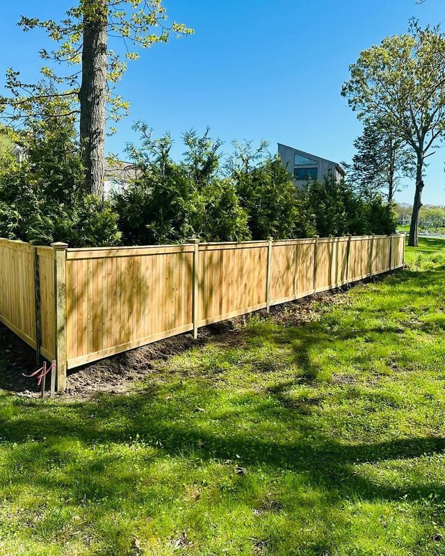 Wooden fenced enclosure in a sunny grassy yard with trees and a house in the background