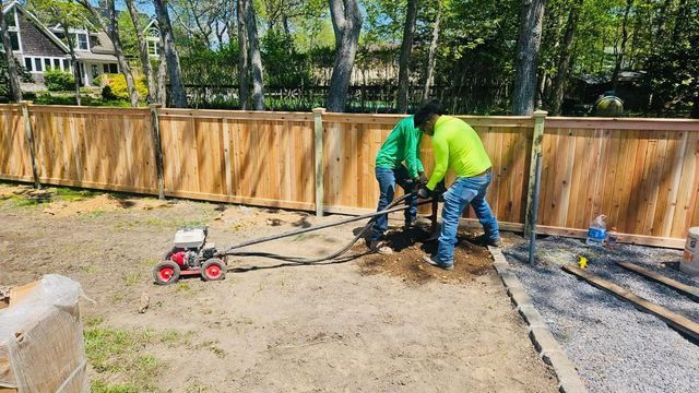 Workers using a sod cutter in a backyard, preparing bare ground along a wooden fence.