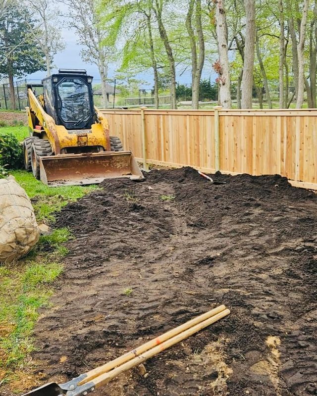 Yellow skid steer leveling dirt beside a wooden fence in a backyard under trees.