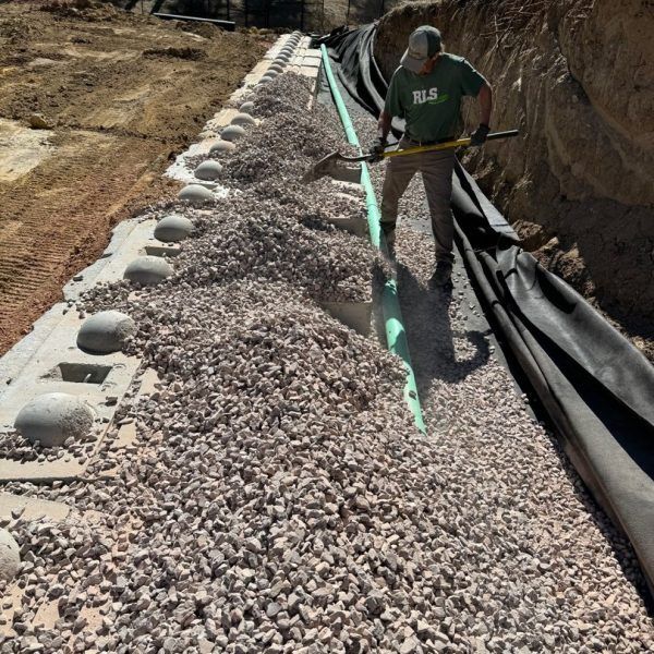 A worker in a green shirt uses a shovel to spread gravel behind a retaining wall with a green drainage pipe.