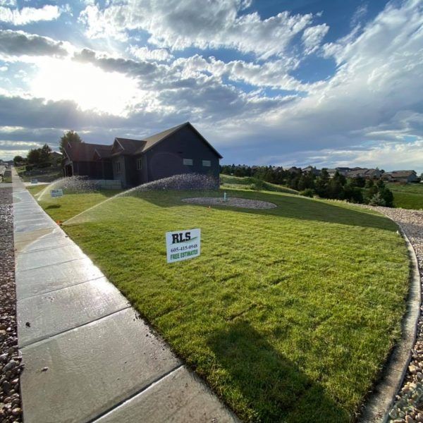 A sunny, landscaped yard with an active sprinkler system, a dark house, and an RLS real estate sign on a bright day.