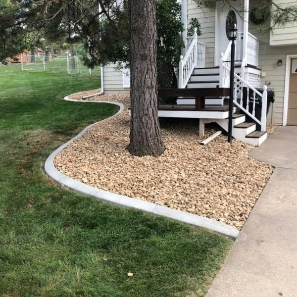 A curved concrete border separates a gravel landscaping bed surrounding a tree from a lawn, leading to a house entrance.