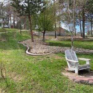 A rustic wooden Adirondack chair sits on a small stone patch in a grassy yard, near a curved stone-lined garden bed.