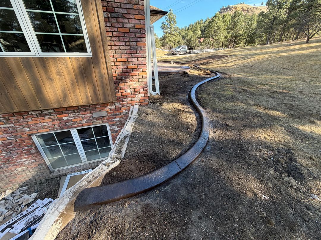 Black drainage pipe curves along the dirt beside a brick house on a sloped yard