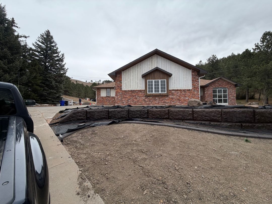 Suburban house with brick and white siding, gravel yard, and stone retaining wall under cloudy sky