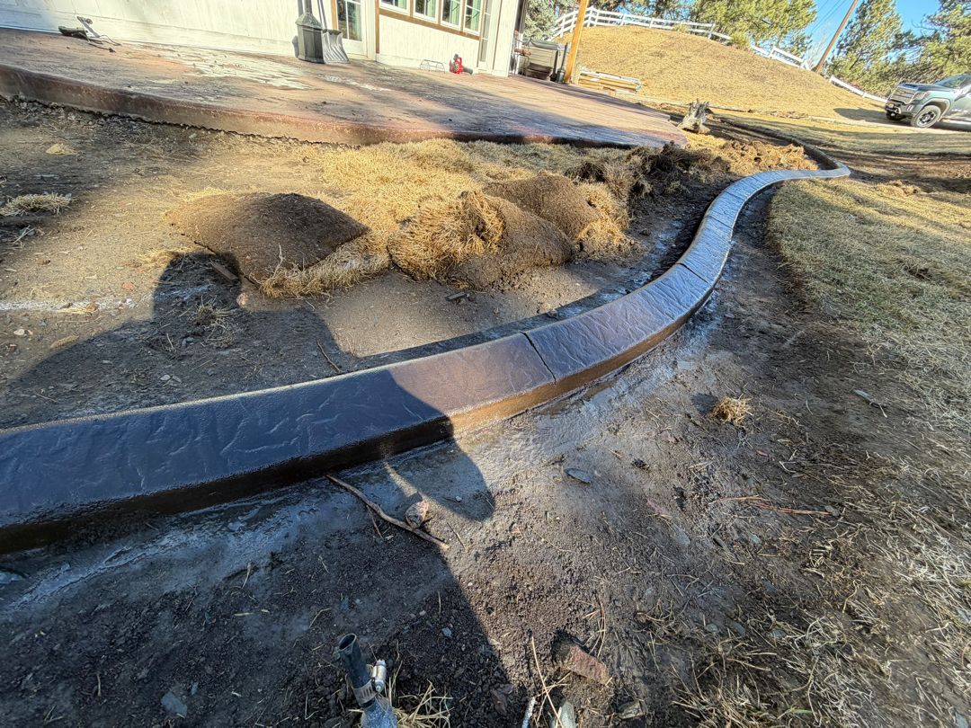 Curved metal strip installed in a dirt trench along a grassy yard near a house, with shadows on the ground