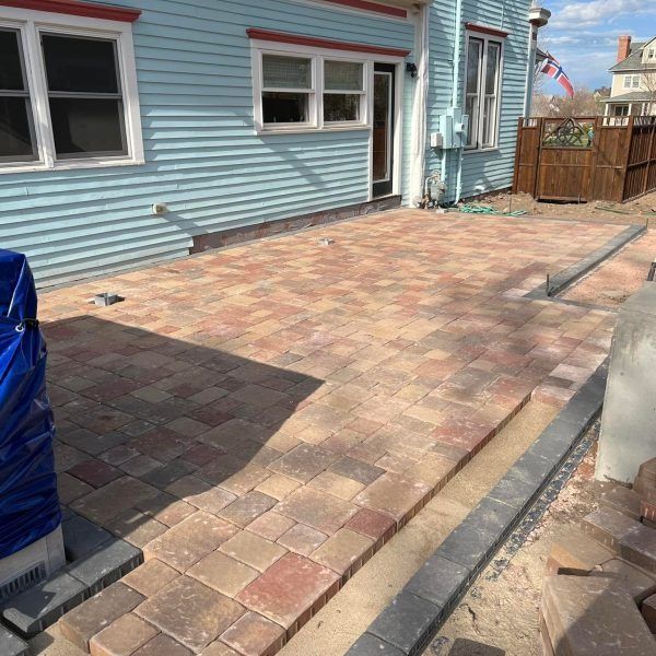 A light blue house exterior with a newly installed multi-colored paver patio and unfinished gray edging along the side.