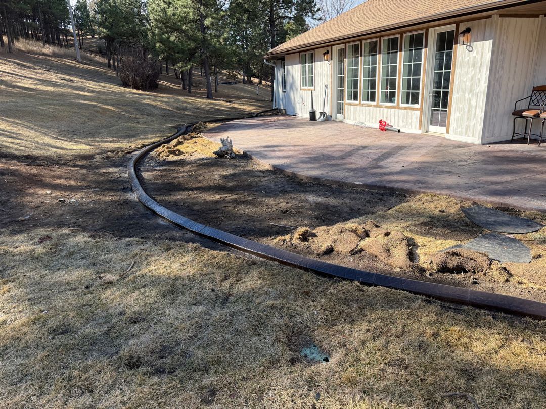 Curved black hose edging a patio beside a beige house on a dry, wooded slope