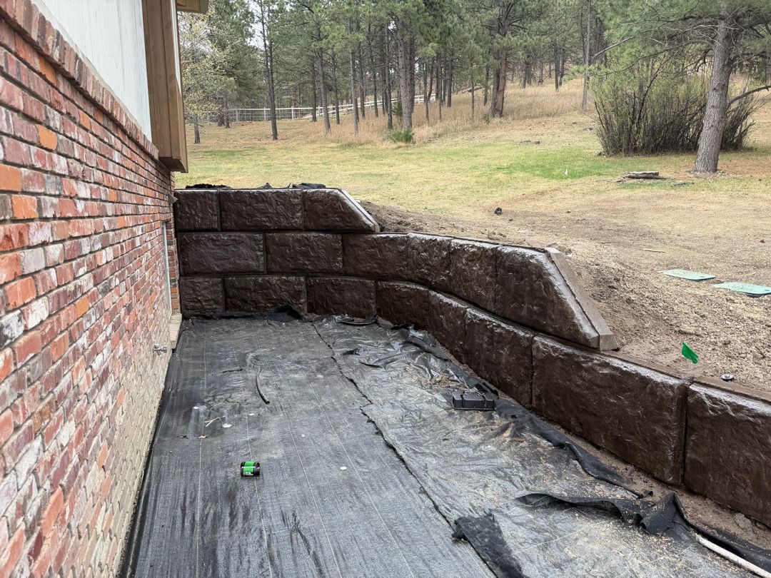 Brick patio beside house with curved stone retaining wall and wooded yard beyond.