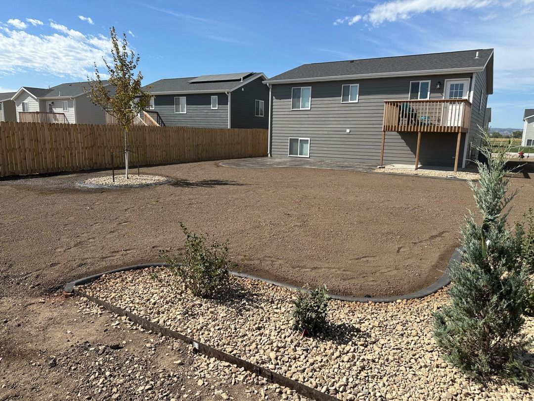 Desert-style backyard with gravel, small shrubs, and a fenced patio beside a gray house under a blue sky