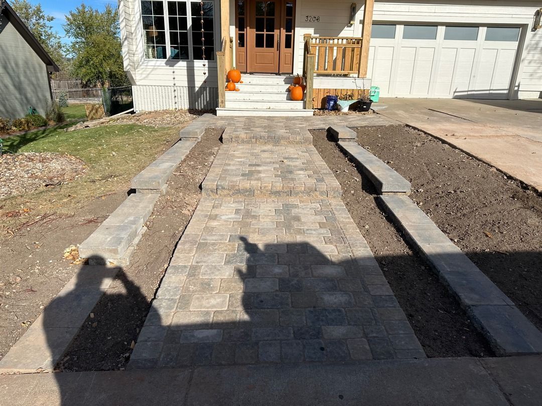 Brick walkway leading to a house entrance with steps, flanked by low concrete borders and garage doors.