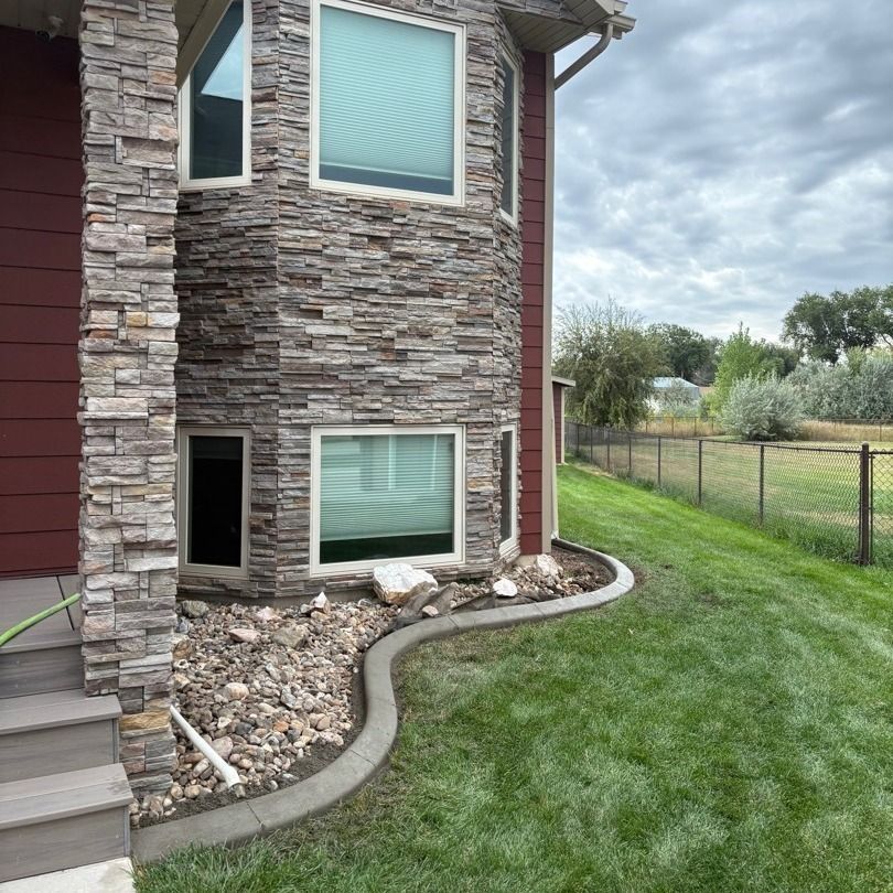 Stone-sided house corner with windows, curved concrete edging, rock bed, and green lawn beside a fenced yard